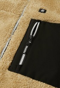 Beige Fleecejacke mit einer schwarzen Stofftasche; ausgestattet mit einem kontrastierenden weißen Reißverschluss und einem Logotag. Weiche Textur und eine behagliche Ausstrahlung.