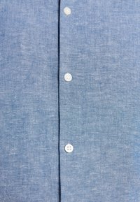 Tissu texturé bleu clair avec trois boutons blancs alignés verticalement au centre, montrant un détail de l'avant d'une chemise.