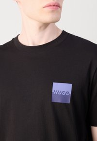 T-shirt en coton noir avec un petit patch logo violet portant le mot "HUGO" sur la zone de la poitrine à gauche. Manches courtes, design classique à col rond.