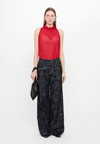 Rode sheer mouwloze top met gerimpelde halslijn; gecombineerd met wijdvallende zwarte broek met bloemenpatronen; houdt een zwarte handtas vast.