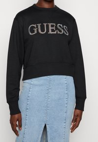 Personne portant un sweat cropped noir avec le logo "GUESS" en strass et une jupe en denim bleu clair taille haute avec une fente devant.