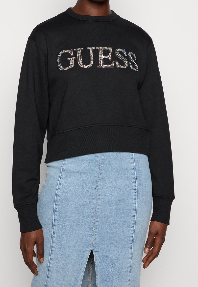 Personne portant un sweat cropped noir avec le logo "GUESS" en strass et une jupe en denim bleu clair taille haute avec une fente devant.