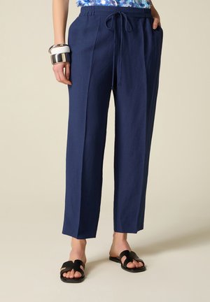 Donna che indossa pantaloni blu navy con coulisse, top blu con fantasia, sandali neri a ciabatta e braccialetti argento e bianchi al polso sinistro.