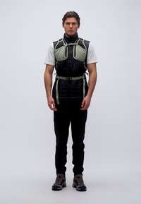 Mees, kes kannab musta tikandiga vestiga heleroheliste rihmade ja taskutega, valget T-särki, musti pükse ja matkajalatsid, seisab ees suunaga.