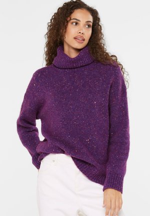 Woman with curly hair wearing a purple turtleneck sweater and white pants, looking slightly to her left with one hand in pocket.