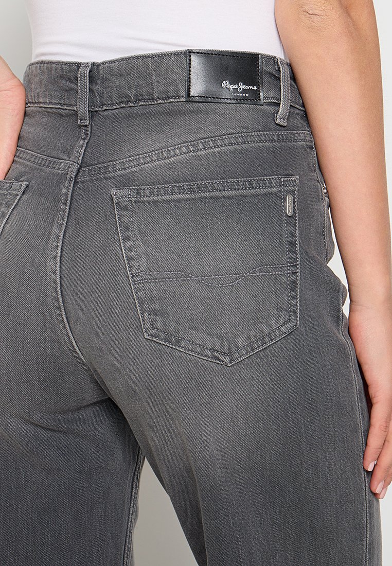 Jean en denim gris à taille haute, avec un patch logo noir à l'arrière, des poches standard et des détails de couture subtils.
