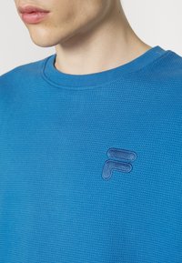 Gros plan d'une personne portant un pull bleu texturé à col rond avec un logo Fila brodé sur la poitrine.