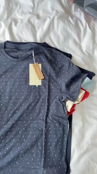 T-shirt azul-marinho com bolinhas brancas, mangas curtas, tecido de algodão suave, inclui uma etiqueta pendurada. Doblado com outras camisetas coloridas.