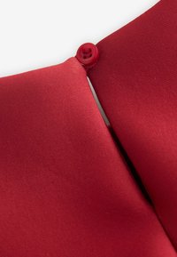 Czerwona tkanina o fakturowanej powierzchni z zapięciem na guzik na szwie. Projekt prezentuje gładki, zakrzywiony kształt w miejscu łączenia się tkaniny.