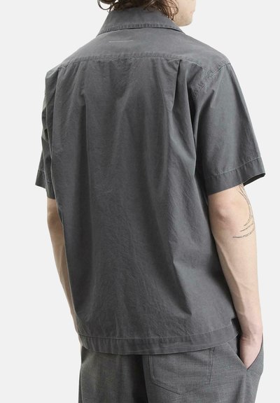 Homme portant une chemise à manches courtes gris foncé et un pantalon gris, debout, une main dans la poche, dos tourné vers la caméra.