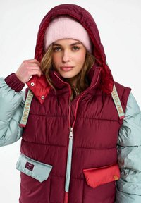 Nő állítja a kapucnit egy ujjatlan, bordó színű, könnyű töltetű mellényen, világoskék ujjakkal és rózsaszín kötött sapkával, egyszínű háttér előtt.