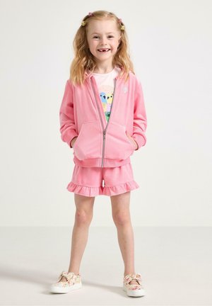 Smilende ung pige med blond hår i to hestehaler iført en pink lynlåshættetrøje, flæse-shorts, skjorte med fugleprint og blomstrede slip-on sko.