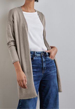 Vrouw draagt een beige openvallend vest over een wit shirt en blauwe spijkerbroek, met één hand in de zak tegen een effen grijze achtergrond.