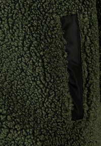 Veste en polaire vert olive avec un extérieur texturé et duveteux et un intérieur en tissu noir lisse. Comprend une poche avec un bord contrastant.