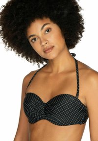 Vrouw met krullend haar die een zwart bikini-topje met kleine witte diamantpatronen en een halternekband draagt tegen een witte achtergrond.