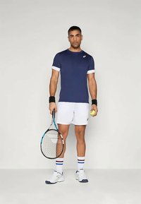 Jūras militārās sporta krekls ar baltām akcéntēm, baltas šorti, melni plaukstu sargi un zili tenis raketes apavi; tur rokās tenis raketi un bumbu.