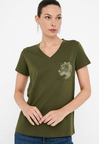 Olive roheline puuvillane T-särk V-kaelusega ja lühikeste varrukatega. Omab taskut, millel on detailne loomagraanul valgetest kivikestest.
