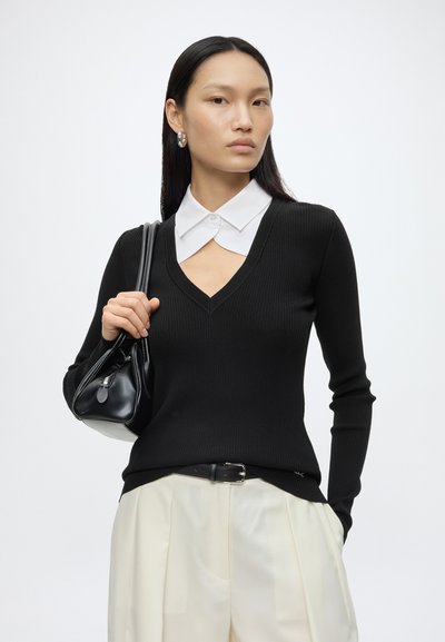 Femme aux longs cheveux lisses, portant un pull noir à col en V sur une chemise blanche à col, tenant un sac à main noir, portant un pantalon blanc plissé.