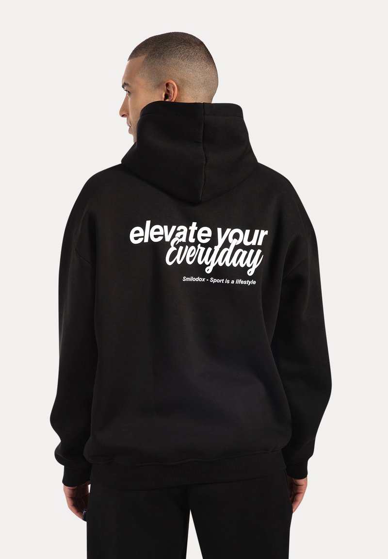 Felpa nera con cappuccio caratterizzata da una grande stampa bianca sul retro che recita "eleva il tuo quotidiano". Il tessuto sembra morbido con una vestibilità comoda e cappuccio.