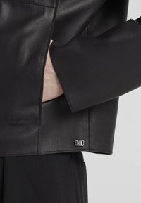 Schwarze Lederjacke mit glatter Textur, die einen seitlichen Schlitz und ein kleines metallisches Logo-Detail nahe dem Saum aufweist.