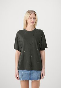 Camiseta oversize verde oscuro de algodón, con tachuelas metálicas dispersas y escote redondo. Combinada con una falda de mezclilla.
