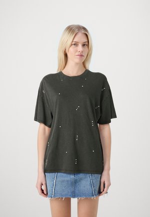 Camiseta oversize verde oscuro de algodón, con tachuelas metálicas dispersas y escote redondo. Combinada con una falda de mezclilla.
