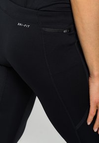 Black Dri-FIT leggings with a side zip pocket, smooth texture, contrasting stitching, and form-fitting design. Material is breathable and stretchy.
