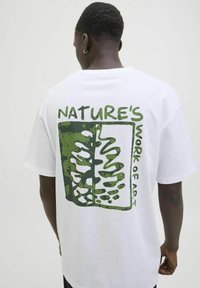 Hvid bomulds t-shirt med et grønt grafisk design på ryggen, der har teksten "NATURE’S WORK OF ART" sammen med en bladillustration.