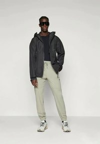 Άνδρας ντυμένος με μαύρη κουκουλωτή ζακέτα, μαύρο πουκάμισο, ανοιχτό γκρι παντελόνι με κορδόνι, λευκά αθλητικά παπούτσια και μεγάλα μαύρα γυαλιά ηλίου, στέκεται σε απλό φόντο.