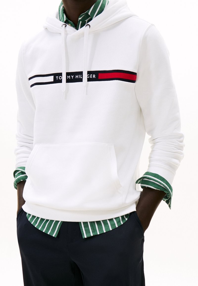 Personne portant un sweat à capuche blanc Tommy Hilfiger par-dessus une chemise rayée verte et blanche, les mains dans les poches d'un pantalon bleu marine.