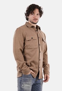 Camicia intrecciata marrone con chiusura a bottoni, due tasche sul petto e colletto. Maniche lunghe e vestibilità rilassata. Indossata con jeans bomber blu chiaro.