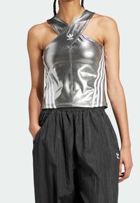 Top corto atlético plateado metálico con tirantes cruzados y logotipo de Adidas en blanco. Combinado con pantalones de pierna ancha texturizados en negro.