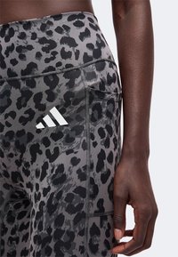 Gros plan d'une personne portant des leggings imprimé léopard gris et noir avec un logo Adidas blanc et une poche latérale, main reposant sur la cuisse.