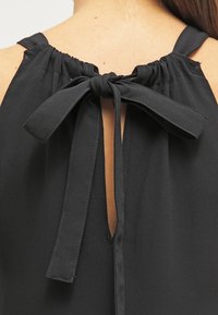 Czarny top bez rękawów z marszczonym dekoltem, wiązaniem z tyłu i wycięciem w kształcie łezki na plecach. Gładka faktura tkaniny.