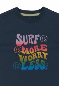 Navy zila kokvilnas T-kreklu ar daudzkrāsainu tekstu, kas lasa "SURF MORE Worry LESS", un uzrakstīts rotaļīgā fontā ar smaidošām sejām.