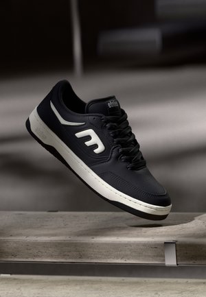 Musta lenkkari, jossa on tekstuuripinta, valkoiset yksityiskohdat ja näkyvä logo sivulla. Varustettu nauhakuormalla ja pehmustetulla nilkkapaneelilla.