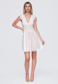 Femme portant une robe en dentelle blanche avec un profond décolleté en V, des manches courtes et une jupe transparente à pois, debout avec une main sur la hanche.