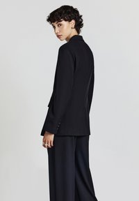 Donna con capelli corti e ricci, che indossa un blazer nero e pantaloni a gamba larga, mentre guarda oltre la sua spalla su uno sfondo semplice.