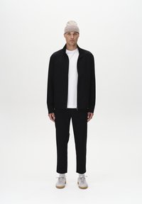 Veste zippée noire avec un col montant, associée à un t-shirt blanc, un pantalon noir et des baskets claires. Le mannequin porte un bonnet en tricot beige.