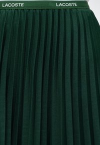 Jupe plissée vert foncé avec taille élastique affichant le texte blanc répété du logo "LACOSTE".
