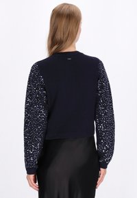 Tengerészsötét pulóver egyszínű testtel és flitteres ujjakkal. Testhez álló fazon, bordás mandzsetta és szegély. Sima textúra fényes részletekkel.