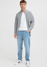 Cárdigan de punto gris claro con un cuello alto, llevado sobre una camiseta blanca, combinado con jeans azul claro y zapatillas blancas con rayas negras.