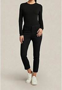 Femme portant un haut noir ajusté à manches longues, un pantalon noir slim et des baskets blanches, debout devant un fond beige uni.