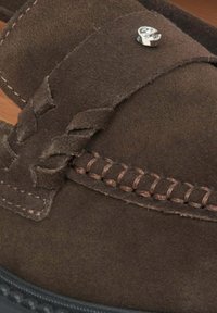 Brown suede slip-on shoe with woven accents and brown stitching. Features a silver decorative element on the strap. Textured surface.