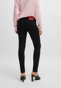 Jeans neri aderenti con vita alta, dotati di un'etichetta rossa con il logo HUGO sul retro, abbinati a un maglione lavorato a maglia rosa chiaro e tacchi neri.