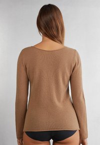 Brauner Langarmpullover aus weichem, strukturiertem Material mit weitem Ausschnitt und leicht ausgestelltem Saum. Dunkle Shorts sind sichtbar.