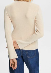 Krémový žebrovaný top s dlouhým rukávem a rozšířenými manžetami, obepnutý střih a měkká textura, kombinovaný s tmavě modrými džíny.