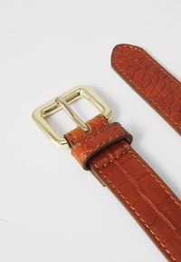 Bruine leren riem met krokodillenpatroon en een gouden metalen gesp. Heeft een gestructureerde oppervlakte en gladde randen. Aangevuld met stikdetails.