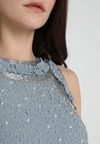 Ljusblå klänning med en volangkant vid halsen, prydd med silverfärgad pärlmönster och pärlaccent, som visar på intrikata detaljer.