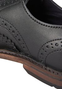 Chaussure en cuir noir avec des perforations brogue détaillées et une semelle en bois. Texture lisse avec des accents cousus le long du bord.